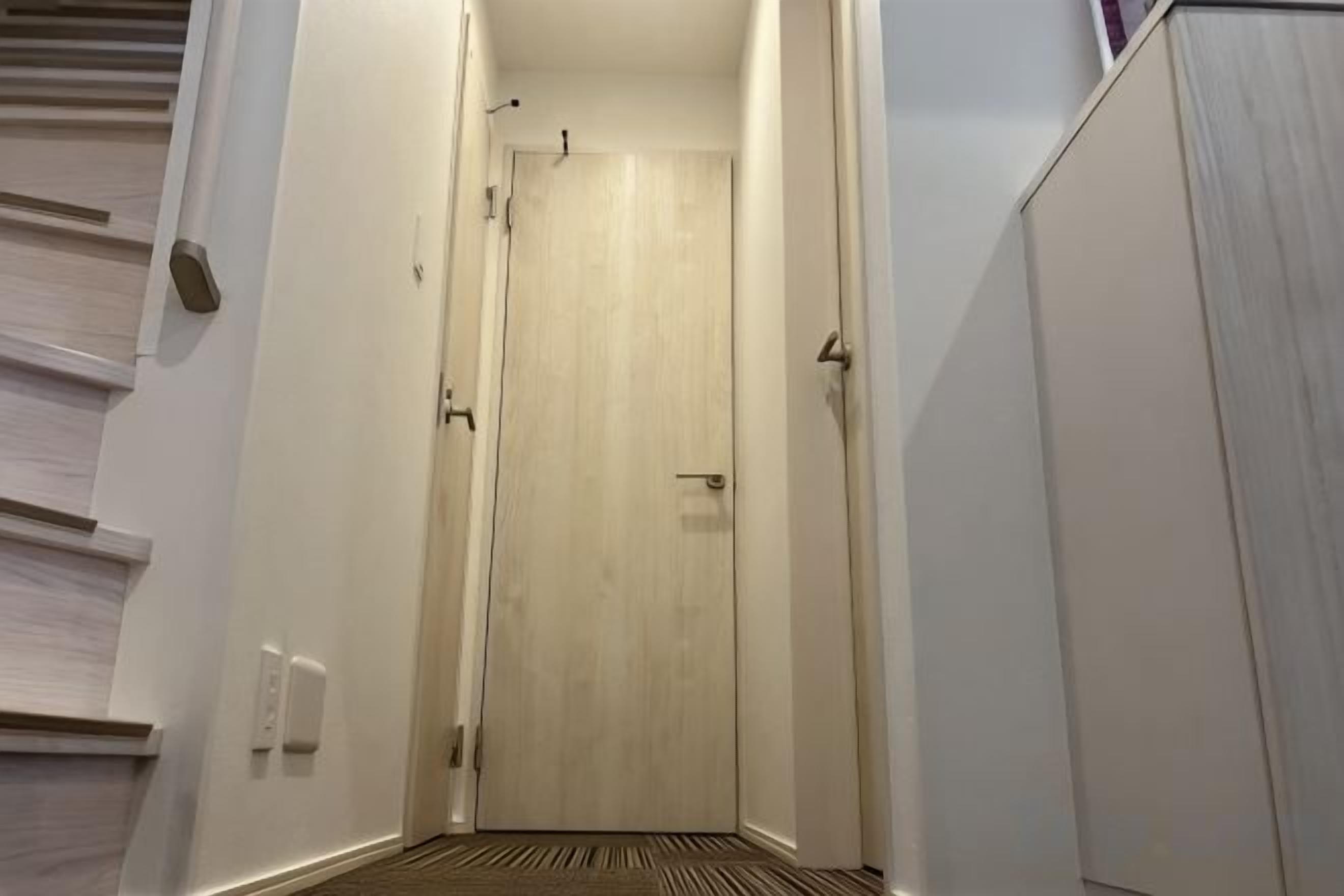 A narrow hallway with a wooden door at the end, a light switch on the wall, and a door handle.