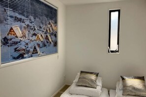 A room with a large window displaying a snowy village scene, a small window, and two beds with pillows. - Four Seasons Yado 1 (Tokyo)