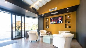 A modern lobby with two white leather sofas, a glass partition, a flat-screen TV mounted on the wall, and a small table with a vase of flowers. - hotel ferrari milano (Carugate)