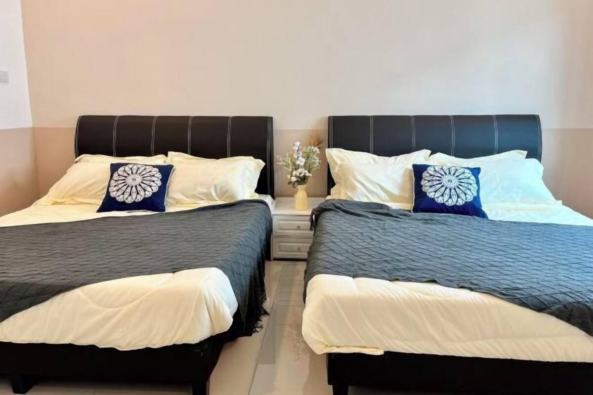 Two twin beds with black headboards and white bedding, a small bedside table with a vase of flowers, and a plain wall in the background.