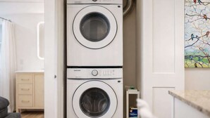 A compact, modern laundry room with two stacked washing machines, a small shelf with a detergent box, and a painting on the wall.