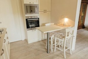 Una cocina compacta con armarios blancos, un microondas, un horno, una mesa de comedor con dos sillas y un ventanal pequeño.