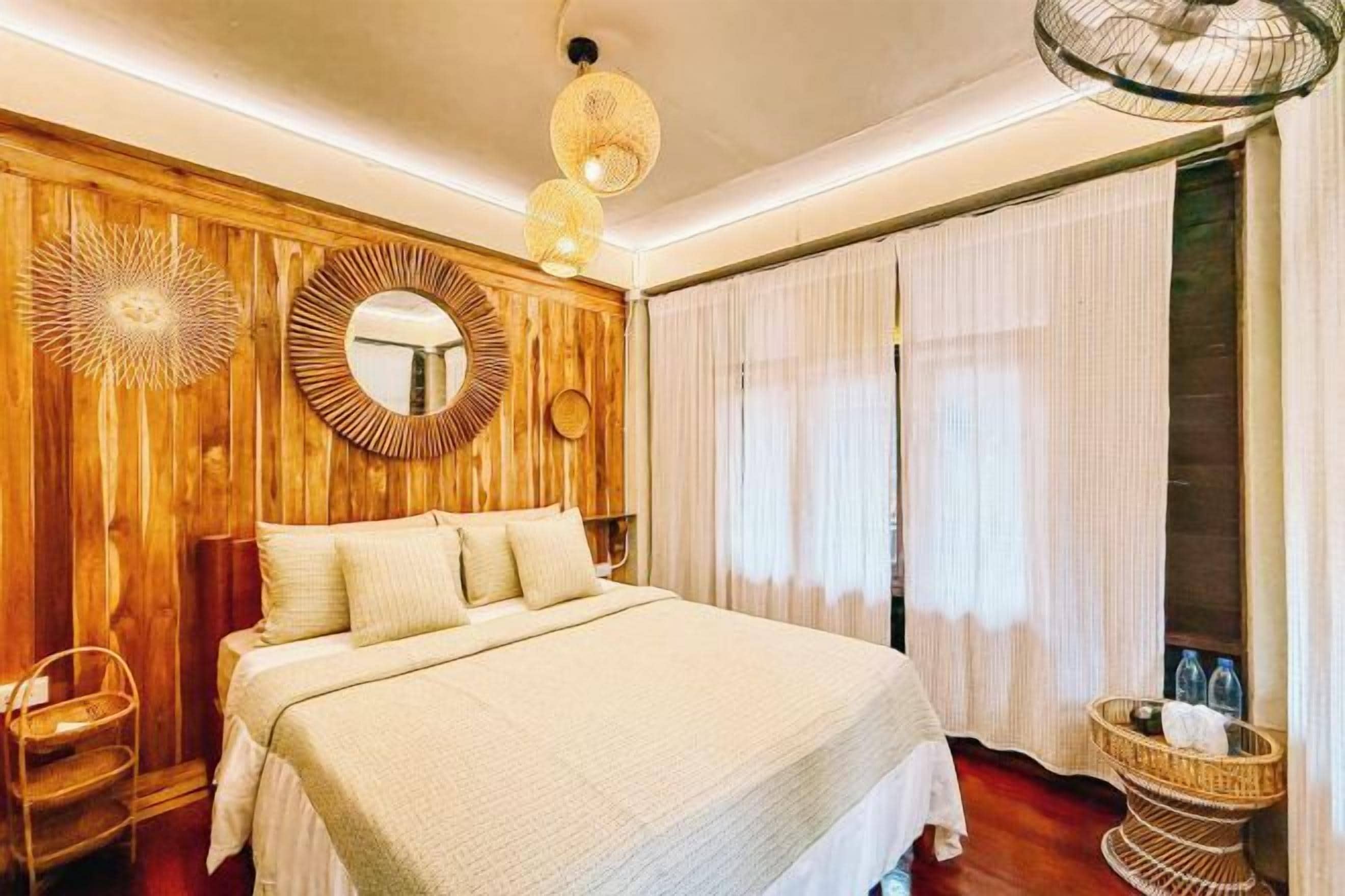 Un dormitorio con una cama grande, paredes de paneles de madera, un espejo redondo y una mesita de mimbre.