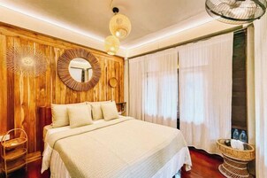 A bedroom with a large bed, wooden panel walls, a round mirror, and a wicker side table.