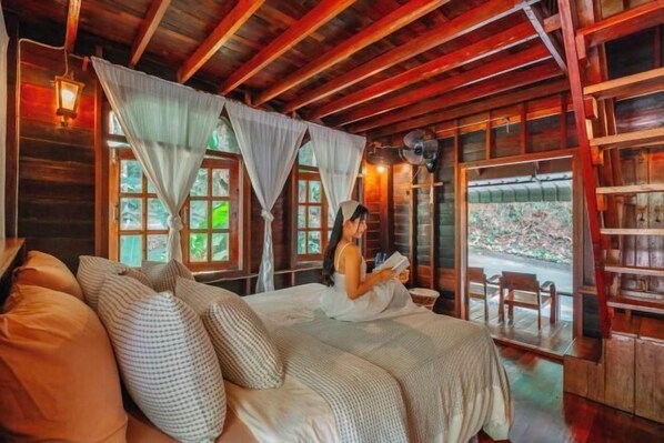 Interior de una cabaña de madera con cama, zona de lectura y grandes ventanales.