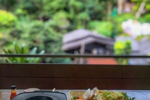 A wooden table with a bottle, a plate, and a bowl of food, with a blurred background of greenery and a building. - Himdoihome Maekampong (Mae On)