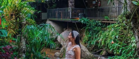 A person sitting by a water feature in a lush garden with a wooden bridge overhead.
