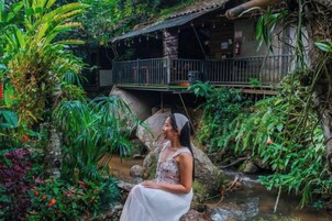 A person sitting by a water feature in a lush garden with a wooden bridge overhead.