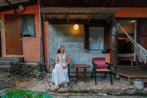 Une personne assise sur une chaise à l’extérieur d’un bâtiment, devant une zone pavée de pierres, avec un hamac et une petite table.