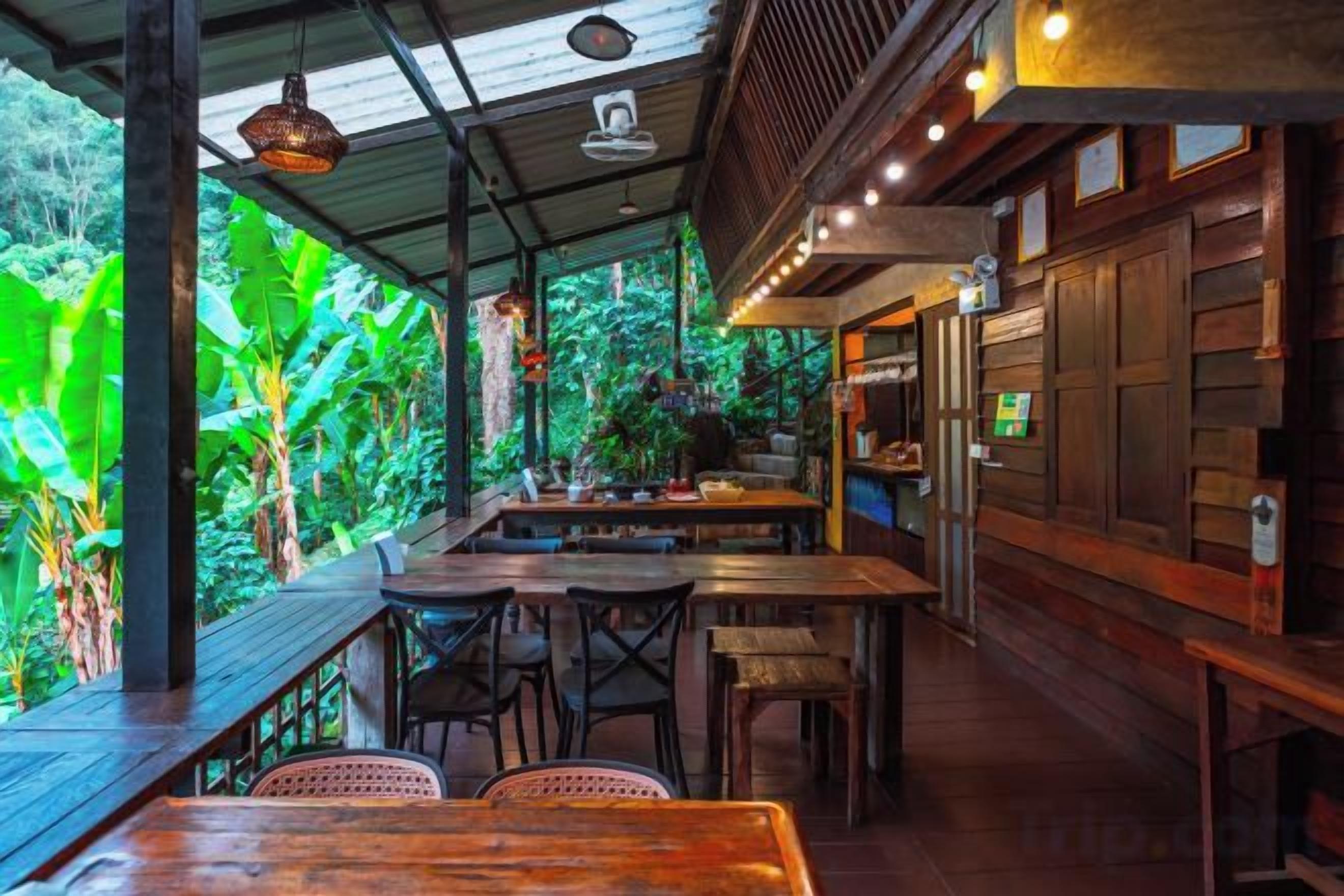 An indoor restaurant with wooden tables and chairs, a counter, and a view of lush greenery outside.
