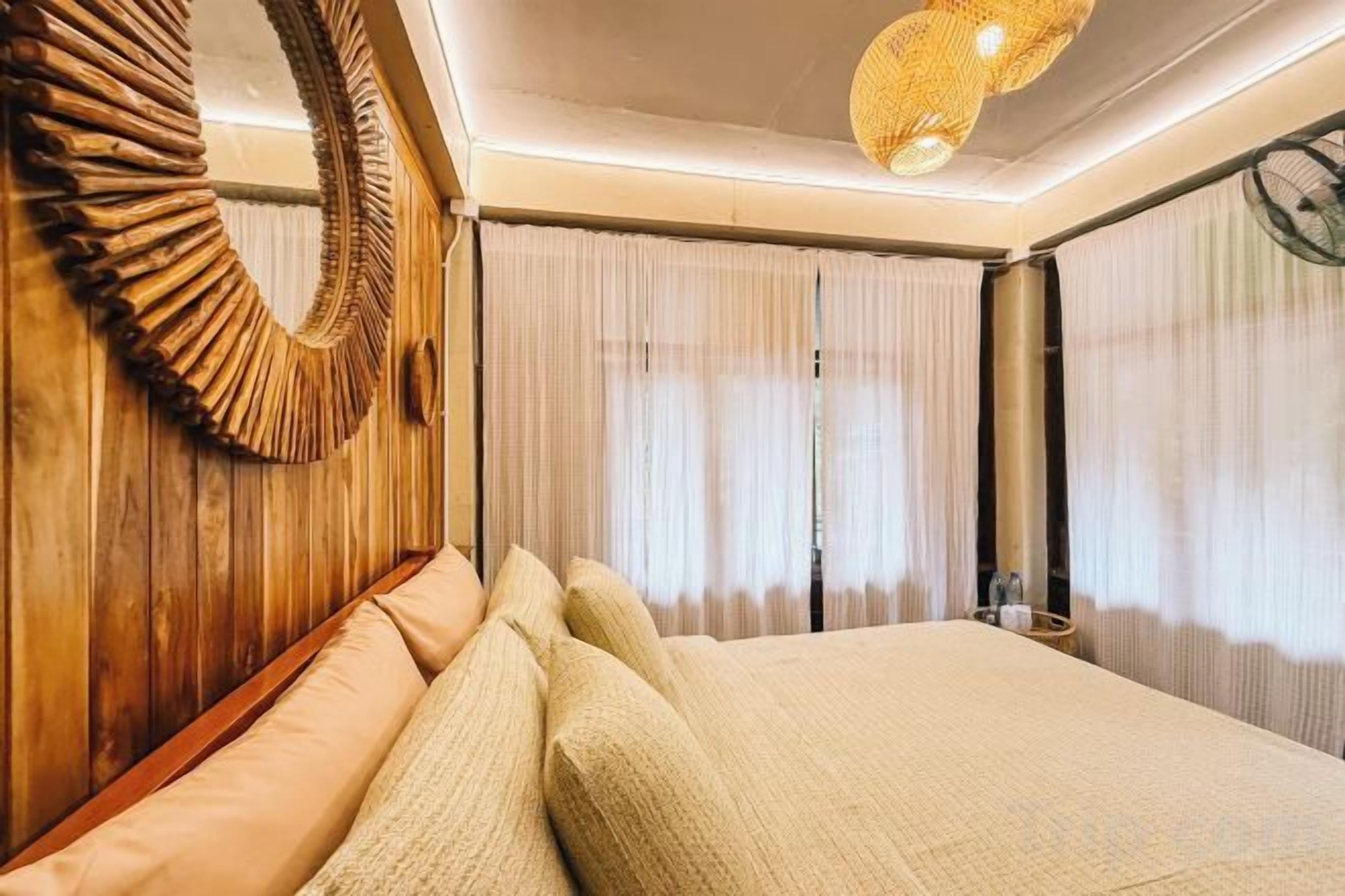 Une chambre d’hôtel avec un grand lit, une tête de lit en bois, un miroir rond et un ventilateur de plafond.
