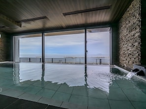 An indoor swimming pool with a view of the sea, stone walls, and a wooden ceiling. - Atami Hotel Nagisakan (Atami)