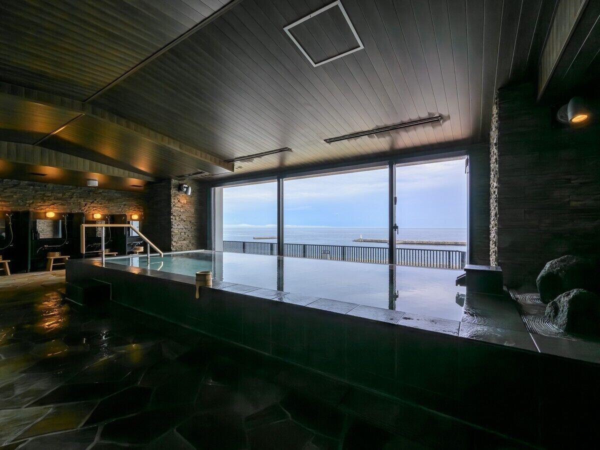 Amplio espacio interior con un gran ventanal con vista a una masa de agua, suelo de piedra y techo de madera.