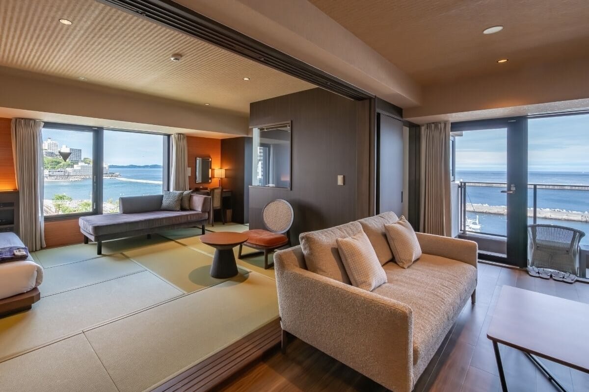 Una sala de estar moderna con un sofá, una mesita y una silla, ventanas amplias con vista al mar y un balcón con barandilla.