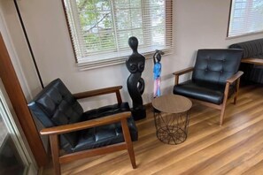 Une pièce comprenant un canapé en cuir noir, un fauteuil en bois, une table basse ronde et une fenêtre avec des stores.