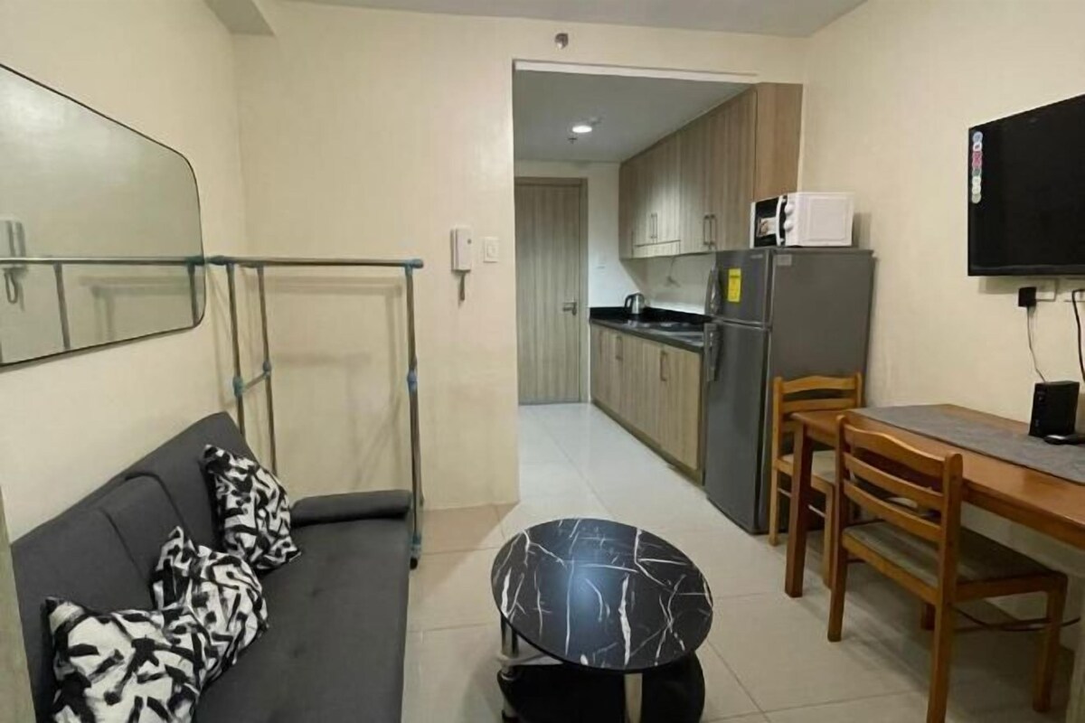 A compact living space with a sofa, a dining table with chairs, a kitchen area, and a television mounted on the wall.