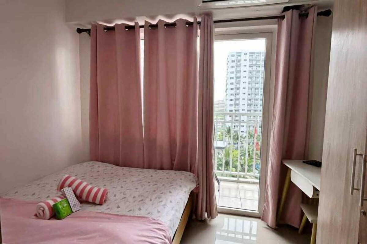 A bedroom with a bed, pink curtains, a window with a view of buildings, and a desk.