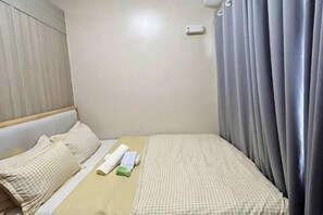 A small, neatly made bed with a checkered bedspread and pillows, a bedside table with a remote control, and a curtain on the window. - MOA getaway staycations (Pasay)