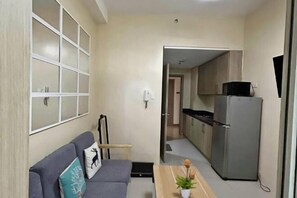 A living room with a sofa, a small table, and a kitchen area with a refrigerator and cabinets. - MOA getaway staycations (Pasay)