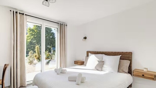 A bedroom with a large bed, a wooden headboard, a nightstand, and a view of greenery through a sliding glass door. - Villa 200m²- swimming pool at the gateway to the Beaujolais region (Sainte-Euphémie)