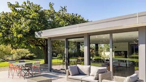 An outdoor patio with wicker furniture, a glass-enclosed area, and a view of a garden. - Villa 200m²- swimming pool at the gateway to the Beaujolais region (Sainte-Euphémie)