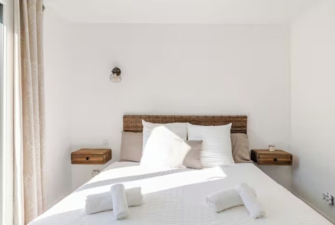 Una cama bien hecha con sábanas y almohadas blancas, cabecera de madera, mesitas de noche con lámparas y una lámpara de pared.