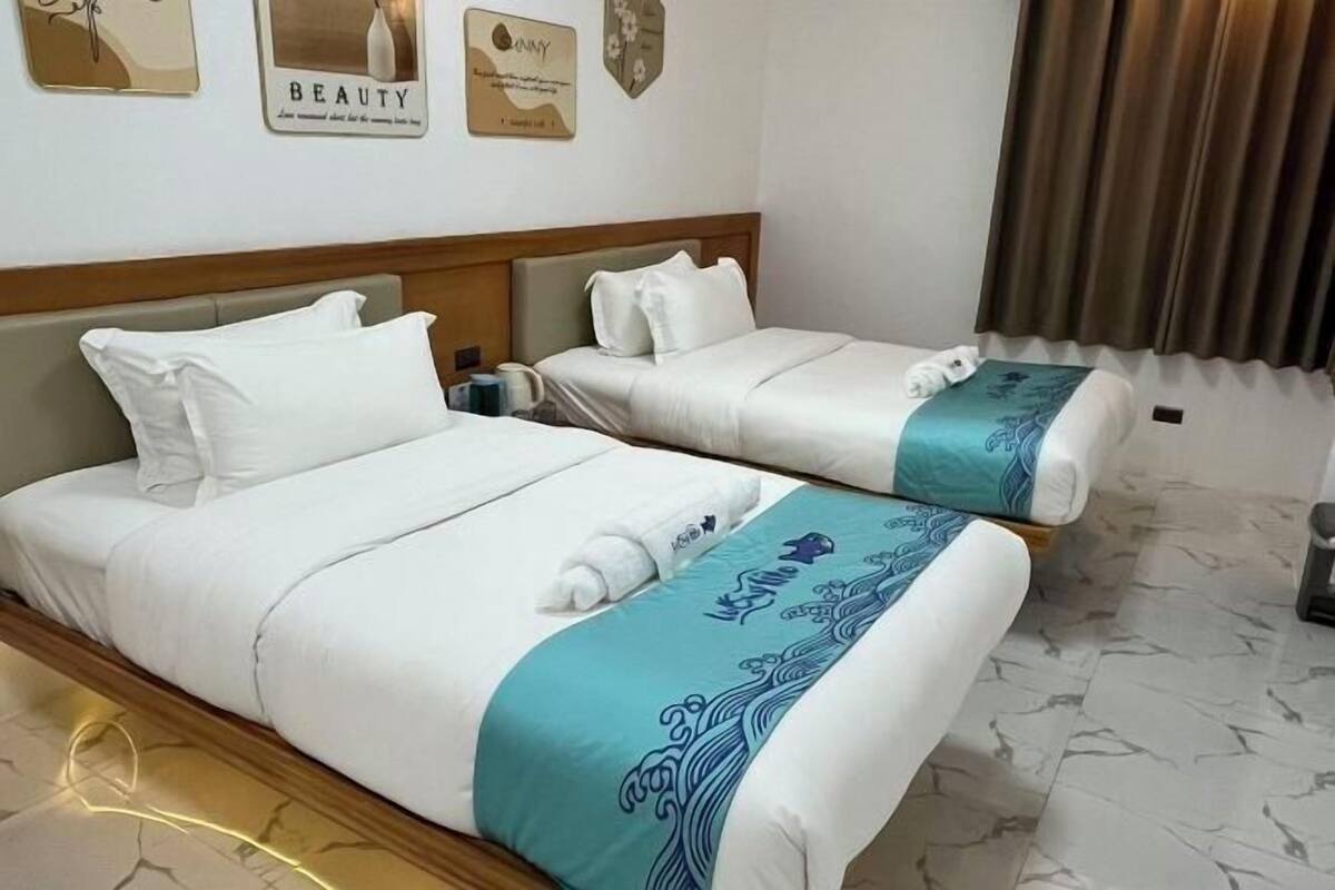 Ein Hotelzimmer mit zwei Betten, jeweils mit weißer Bettwäsche und einem blauen Zierkissen. An den Wänden sind Kunstwerke zu sehen, außerdem ein Vorhang.
