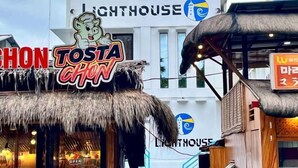 Un ristorante chiamato 'Tosta Chon' con tetto di paglia, situato di fronte a un edificio con un cartello che recita 'Lighthouse'.