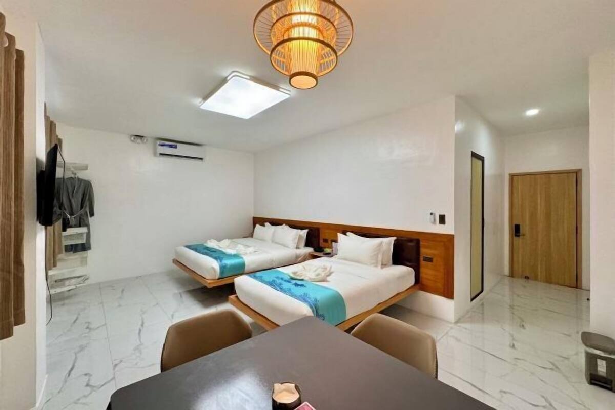 Ein Hotelzimmer mit zwei Betten, einem Esstisch, einer Deckenleuchte, einer Klimaanlage und einer Holztür.
