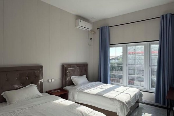 A hotel room with two beds, an air conditioning unit, a window with curtains, and a view of the outside.