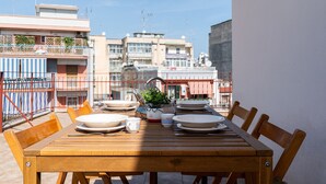 Un tavolo da pranzo in legno apparecchiato con piatti, tazze e una teiera, circondato da sedie, con edifici sullo sfondo.