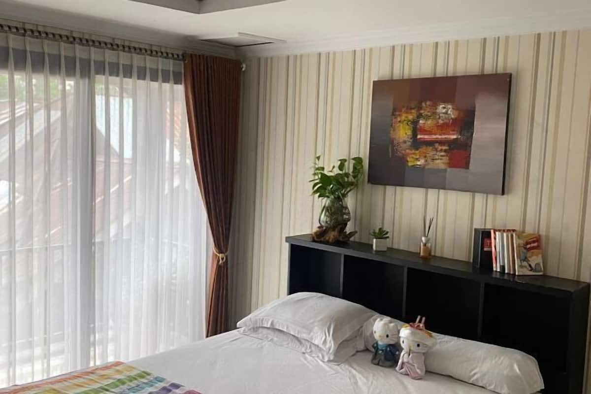 Une chambre d’hôtel avec un lit, une console sombre sur laquelle sont posés des livres et une plante, un tableau au mur et une fenêtre avec des rideaux.