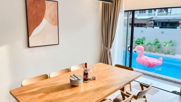 A dining area with a wooden table and chairs, a framed artwork on the wall, and a view of a swimming pool with a pink flamingo float. - HH Villa Private Pool Bercham (Ipoh)