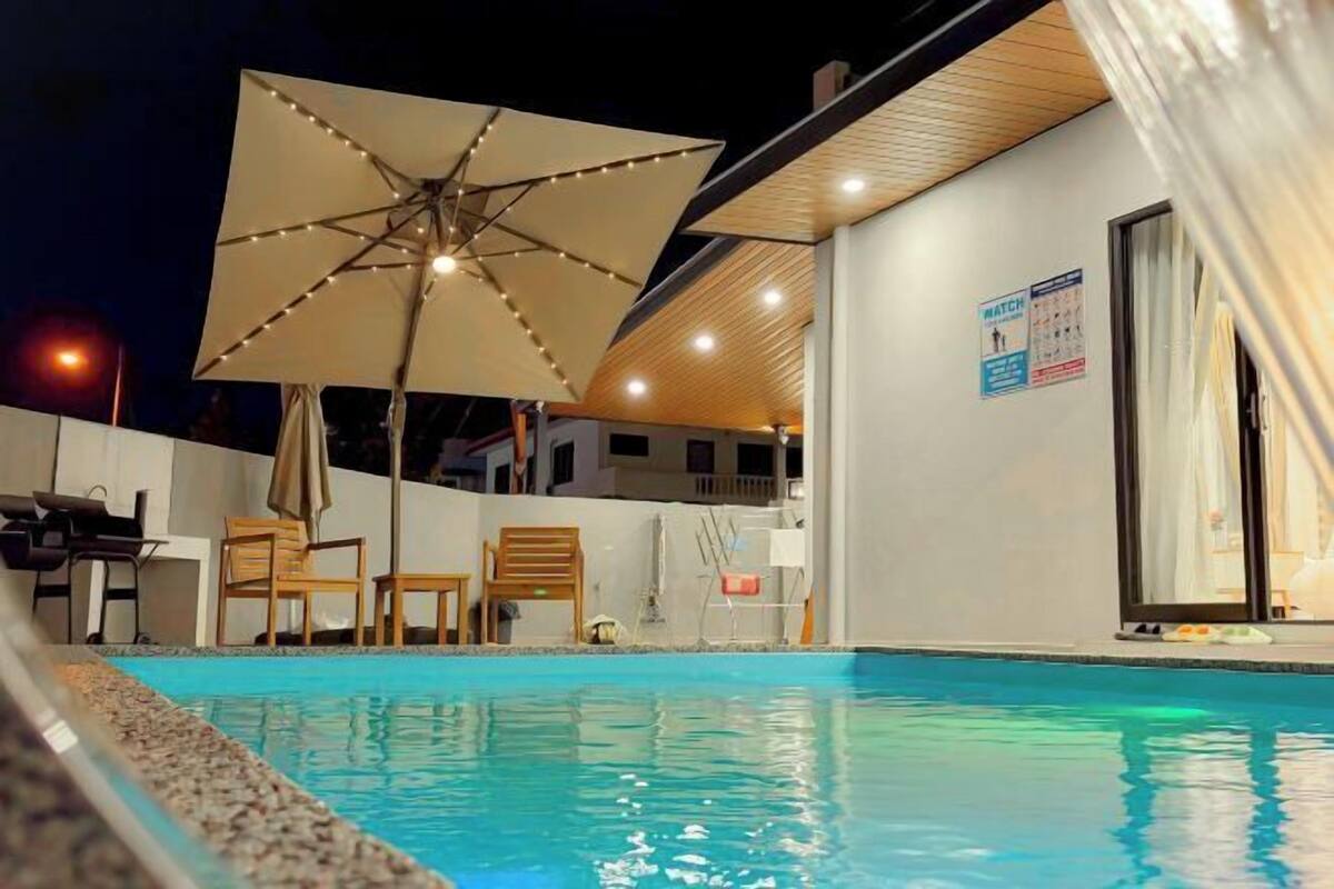 A swimming pool with lounge chairs and an umbrella, a sign with a person icon, and a building with a wooden ceiling.