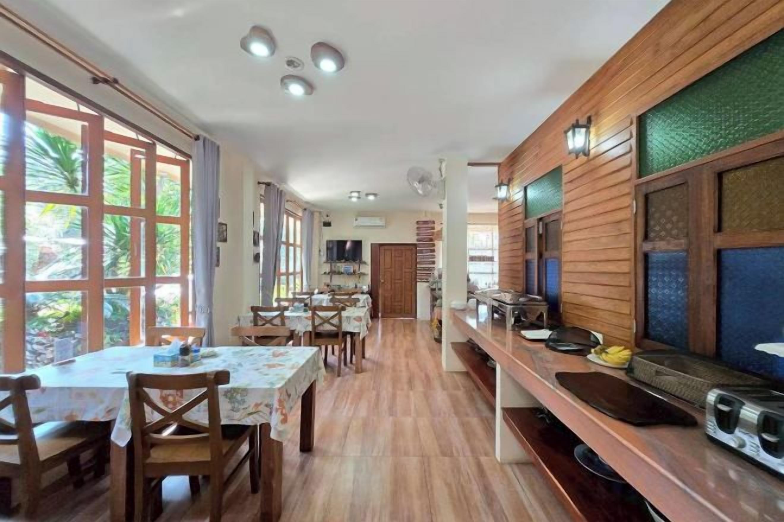 A dining area with wooden tables and chairs, a long counter with kitchen appliances, and large windows with curtains.