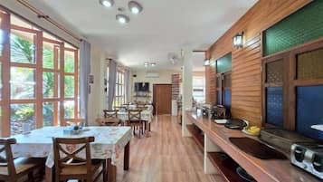 A dining area with wooden tables and chairs, a long counter with kitchen appliances, and large windows with curtains.