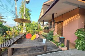 A wooden deck with a bench and a sign, a building with a sloped roof, and a large plant. - Baan Yung Hill Resort (Chiang Rai)