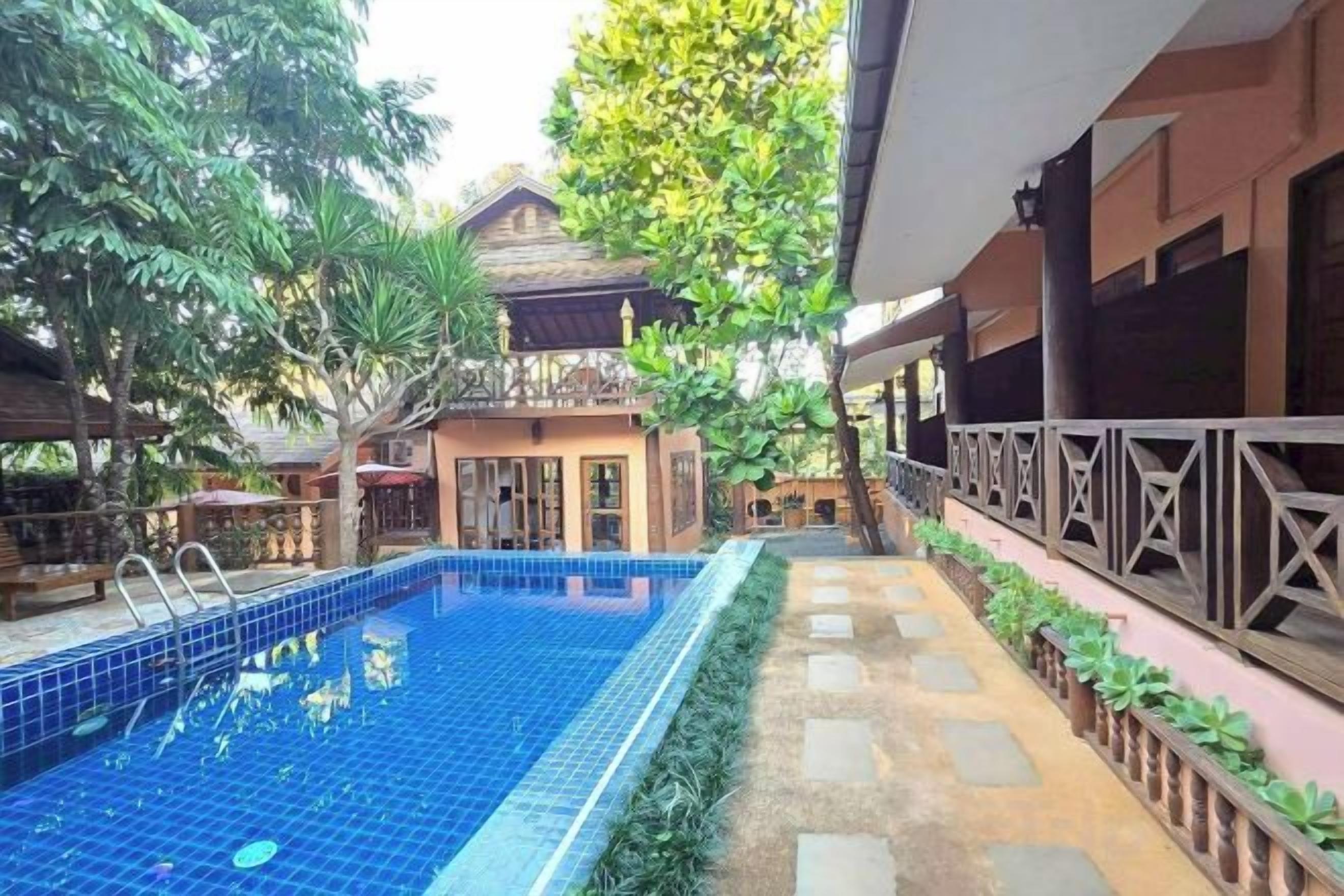 A swimming pool with a tiled deck, surrounded by lush greenery and a building with a balcony.