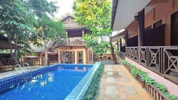 A swimming pool with a tiled deck, surrounded by lush greenery and a building with a balcony.