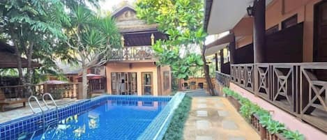A swimming pool with a tiled deck, surrounded by lush greenery and a building with a balcony.