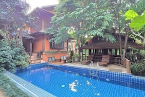 A swimming pool with blue tiles, surrounded by greenery and a wooden deck with chairs.