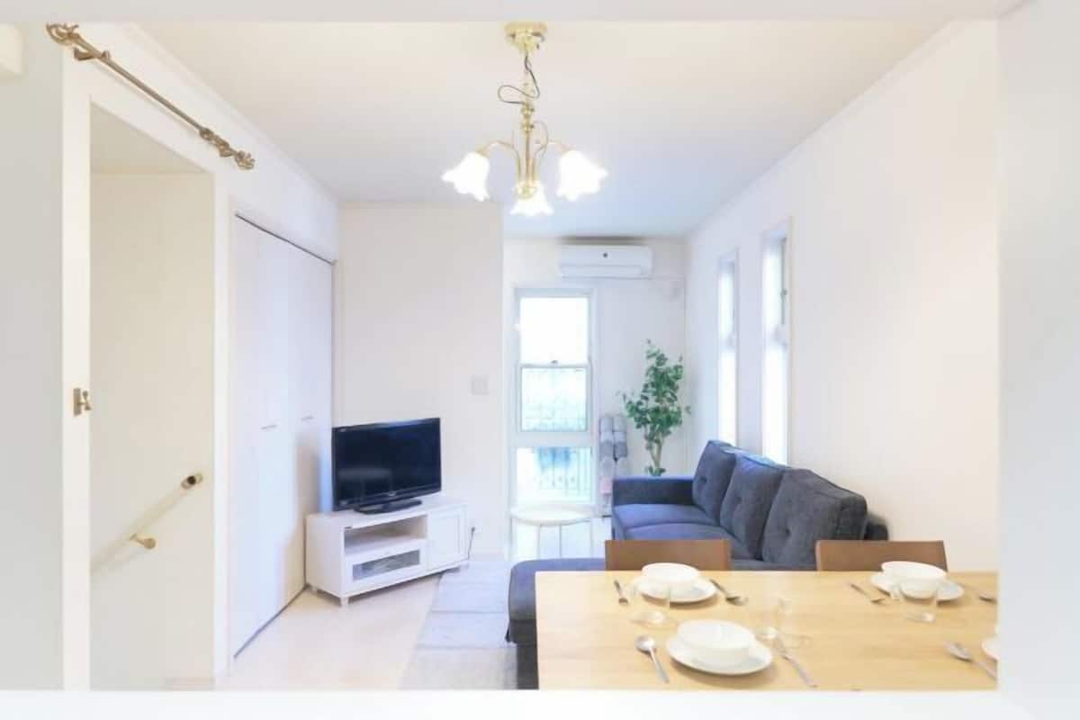 Una sala de estar moderna con un sofá, una mesa de comedor para dos personas, un televisor sobre un mueble bajo y una araña.