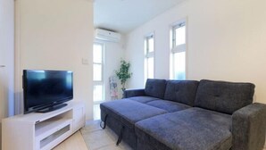 A living room with a flat-screen TV on a white stand, a grey sectional sofa, and a potted plant. - Etoile Soshigaya Okura by Tranova (Tokyo)