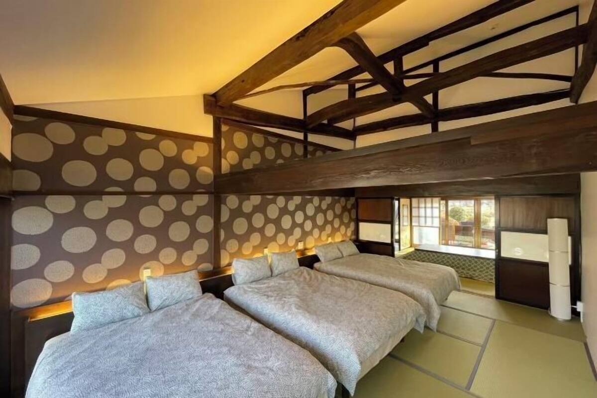A room with a high ceiling, exposed wooden beams, and a traditional Japanese tatami mat floor. There are three beds with grey bedding and a large window providing natural light.