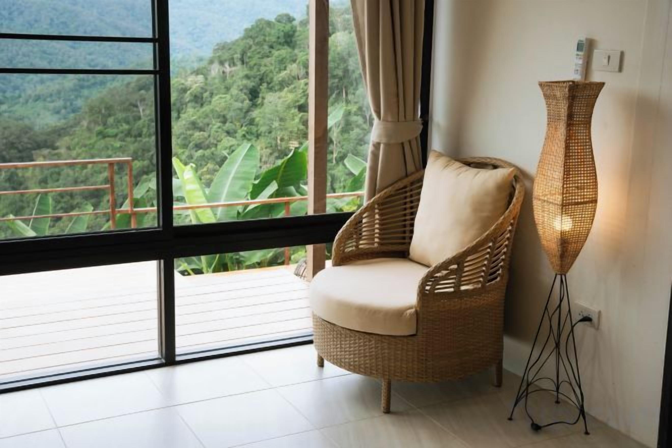 Una silla de mimbre con cojín, una lámpara de pie alta y un ventanal con vistas a la vegetación.