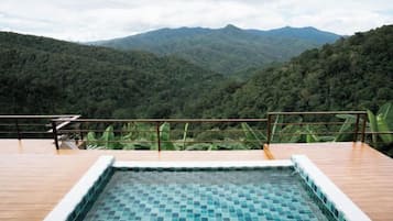 A rectangular pool with mosaic tiles, surrounded by a wooden deck and a metal railing, overlooking a lush forested mountain range.