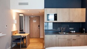 A compact hotel room with a wooden floor, a bed, a small dining table with chairs, a kitchenette with a microwave, and a door leading to another room. - Holiday Inn Express & Suites MONHEIM AM RHEIN by IHG (Bedburg)
