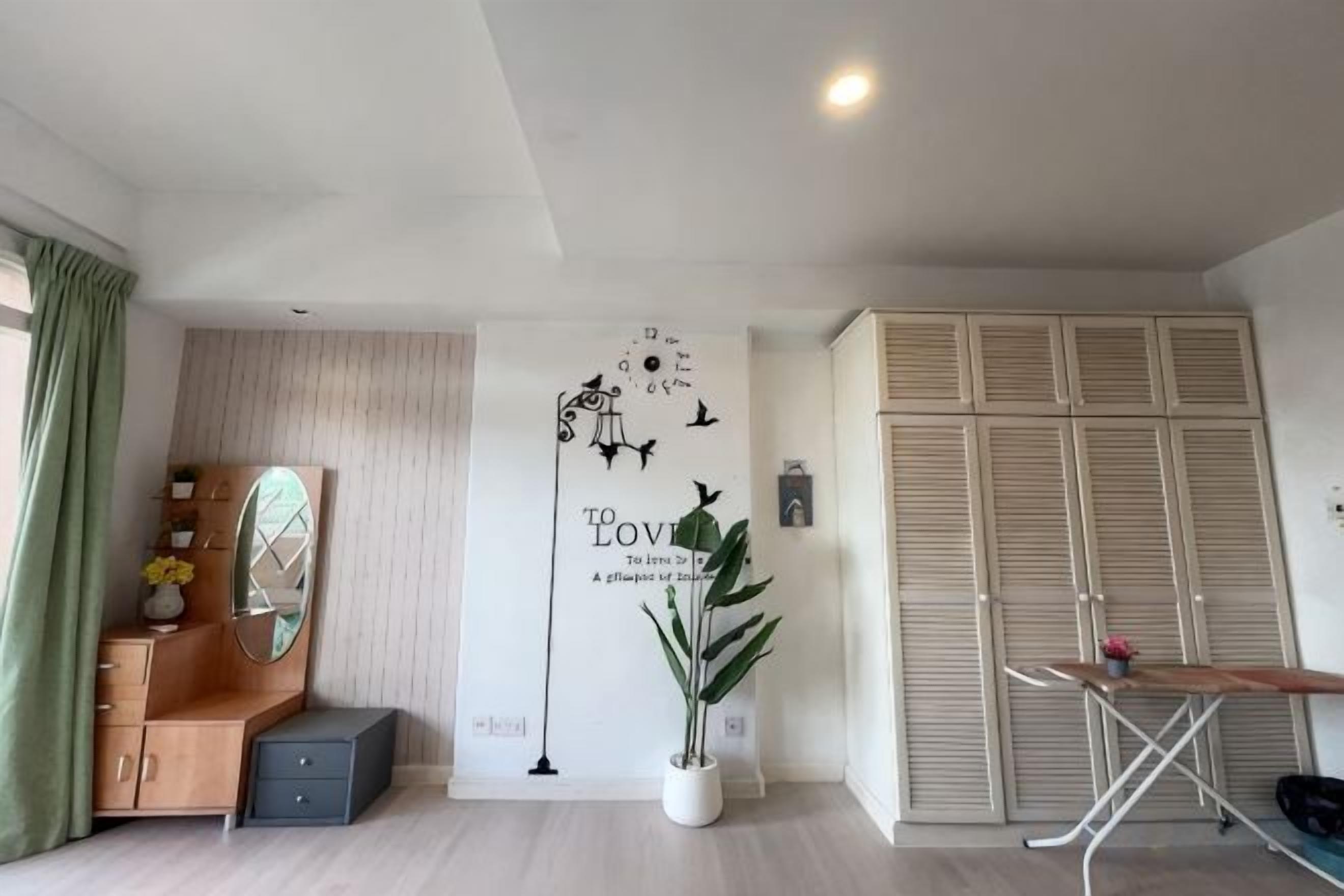 A room with a wooden dresser, a mirror, a potted plant, a wall with decorative text, and a closet with louvered doors.