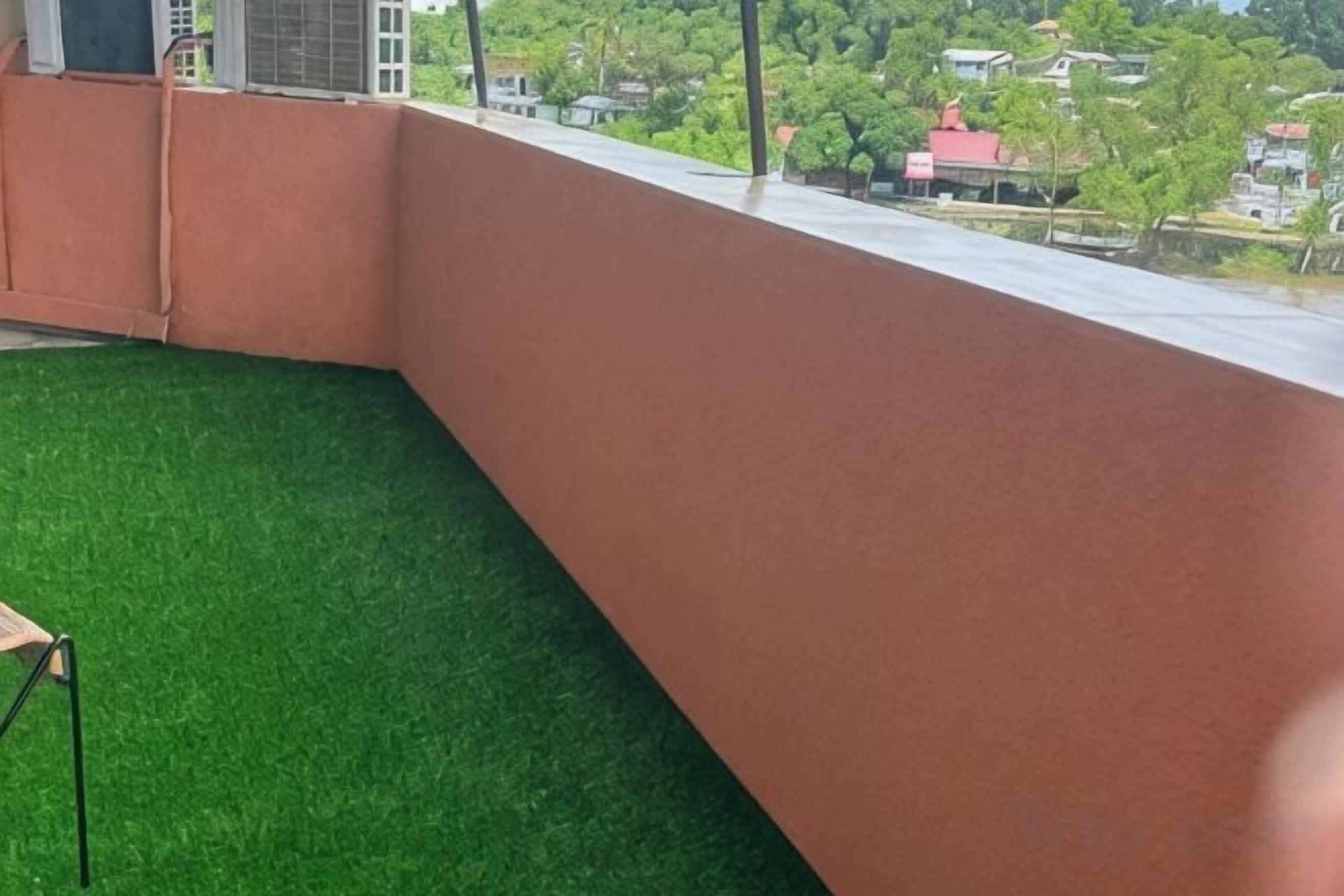 A balcony with artificial grass flooring, a metal chair, and a view of a residential area with houses and trees.
