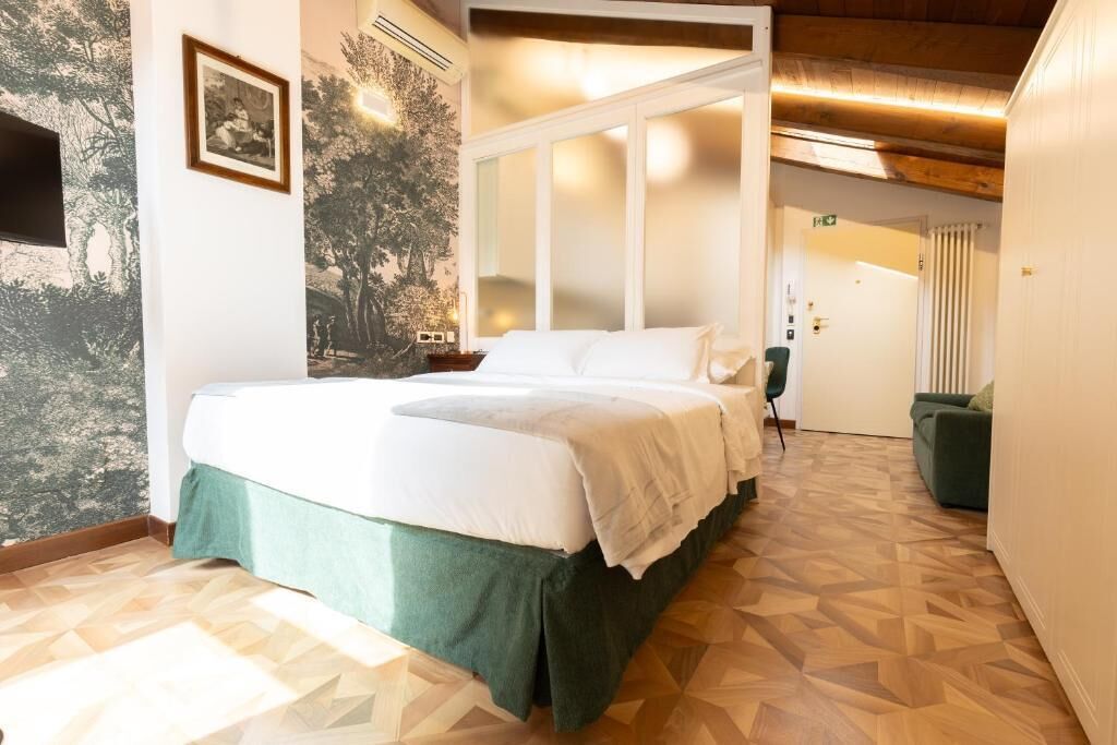 Une chambre d’hôtel avec un lit, un couvre-lit vert, un mur orné d’une fresque représentant un arbre, un plancher en bois et une porte donnant sur une autre pièce.